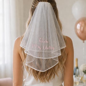 May include: A white two-tier tulle wedding veil featuring a finished edge and pink embroidered text that reads Future Mrs. Cherry. The veil is gathered at the top and drapes down the back, showcasing a soft, sheer fabric with a delicate texture.