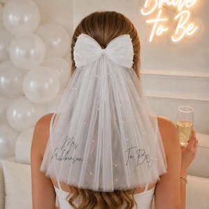 May include: A white tulle wedding veil featuring a large bow at the top, scattered pearl embellishments, and black cursive text reading Mrs. Wilkinson and To Be. The veil is worn over long brown hair, with a neon sign in the background.