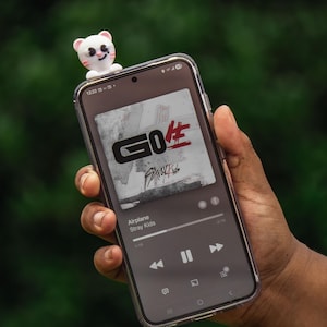 Puede incluir: Un smartphone muestra la interfaz de un reproductor de m&uacute;sica con la car&aacute;tula del &aacute;lbum "GO生" de Stray Kids. Una peque&ntilde;a figura de oso blanco y rosa est&aacute; colocada encima del tel&eacute;fono. La canci&oacute;n "Airplane" se est&aacute; reproduciendo. El tel&eacute;fono se sostiene en una mano.