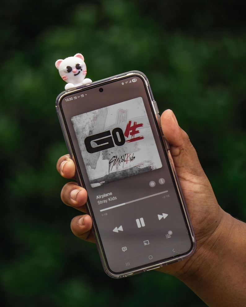 Puede incluir: Un smartphone muestra la interfaz de un reproductor de m&uacute;sica con la car&aacute;tula del &aacute;lbum "GO生" de Stray Kids. Una peque&ntilde;a figura de oso blanco y rosa est&aacute; colocada encima del tel&eacute;fono. La canci&oacute;n "Airplane" se est&aacute; reproduciendo. El tel&eacute;fono se sostiene en una mano.