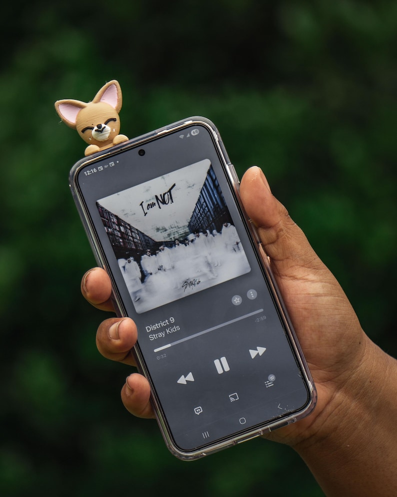 Puede incluir: Un smartphone con una funda transparente se sostiene en una mano. El tel&eacute;fono muestra una aplicaci&oacute;n de m&uacute;sica con la portada del &aacute;lbum "I am NOT" de Stray Kids. Una peque&ntilde;a figura de chihuahua marr&oacute;n claro est&aacute; unida a la parte superior del tel&eacute;fono.