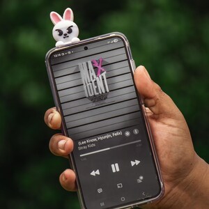Puede incluir: Un tel&eacute;fono inteligente con una funda transparente muestra una interfaz de aplicaci&oacute;n de m&uacute;sica con el texto "MAXIDENT" y "Stray Kids". Una figura de conejo blanco y rosa se encuentra encima del tel&eacute;fono. La pantalla muestra los controles de la canci&oacute;n y la informaci&oacute;n del artista.