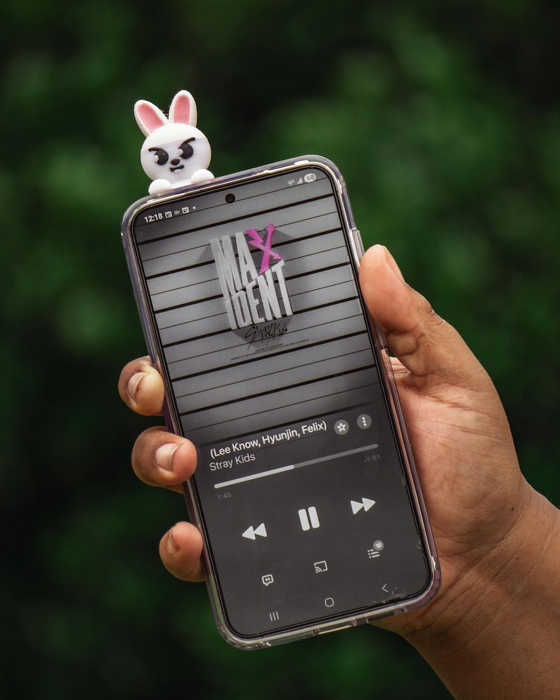 Puede incluir: Un tel&eacute;fono inteligente con una funda transparente muestra una interfaz de aplicaci&oacute;n de m&uacute;sica con el texto "MAXIDENT" y "Stray Kids". Una figura de conejo blanco y rosa se encuentra encima del tel&eacute;fono. La pantalla muestra los controles de la canci&oacute;n y la informaci&oacute;n del artista.