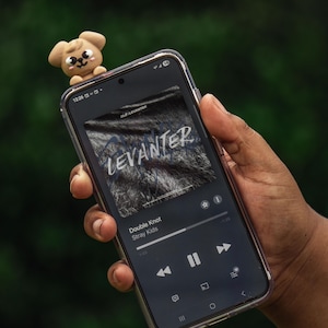 Puede incluir: Un smartphone muestra la car&aacute;tula del &aacute;lbum "Levanter" de Stray Kids, con la canci&oacute;n "Double Knot" sonando. Una peque&ntilde;a figura de perro marr&oacute;n claro est&aacute; encima del tel&eacute;fono. El fondo es verde borroso.