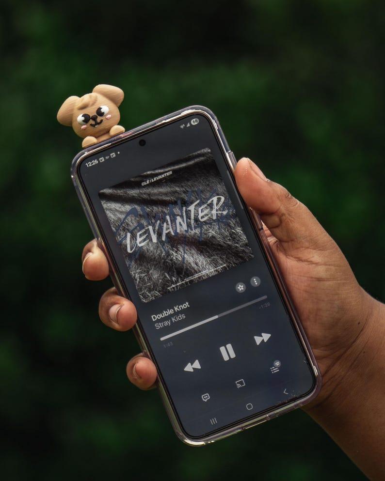 Puede incluir: Un smartphone muestra la car&aacute;tula del &aacute;lbum "Levanter" de Stray Kids, con la canci&oacute;n "Double Knot" sonando. Una peque&ntilde;a figura de perro marr&oacute;n claro est&aacute; encima del tel&eacute;fono. El fondo es verde borroso.