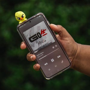 Puede incluir: Un smartphone que muestra la car&aacute;tula del &aacute;lbum "GO生" de Stray Kids, con una peque&ntilde;a figura de pollito amarillo encima. La car&aacute;tula del &aacute;lbum presenta el texto "GO生" en negro y rojo. El tel&eacute;fono se sostiene contra un fondo verde borroso.