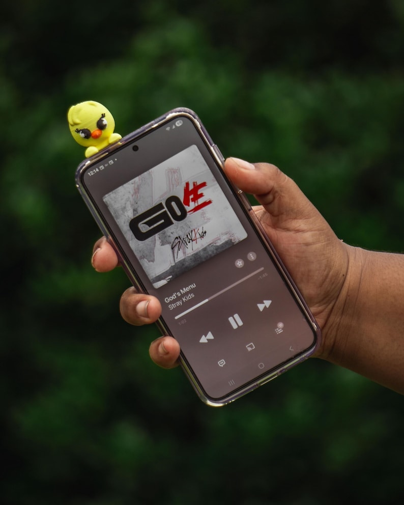 Puede incluir: Un smartphone que muestra la car&aacute;tula del &aacute;lbum "GO生" de Stray Kids, con una peque&ntilde;a figura de pollito amarillo encima. La car&aacute;tula del &aacute;lbum presenta el texto "GO生" en negro y rojo. El tel&eacute;fono se sostiene contra un fondo verde borroso.