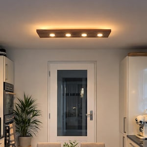 Può includere: Una cucina moderna con un tavolo da pranzo in legno e sei sedie imbottite grigie. Un apparecchio di illuminazione in legno con quattro faretti a incasso è appeso sopra il tavolo. Una pianta in una ciotola di legno è sul tavolo.