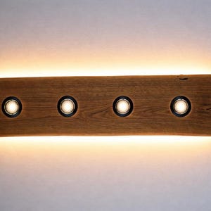 May include: A rectangular wooden wall light fixture with four recessed lights. The wood has a natural grain and a warm brown tone. The lights are circular with black trim, emitting a soft, warm glow. The fixture is mounted on a white wall.