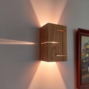 May include: A wooden wall sconce with horizontal slits that emit light. The sconce is rectangular and made of light-colored wood. The light shines onto the wall, creating a warm glow. A framed painting of flowers is next to the sconce.