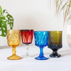 May include: Four colorful vintage wine glasses. The glasses are in shades of amber, red, blue, and olive green. Each glass has a unique design and sits on a stem. The glasses are arranged on a white surface.