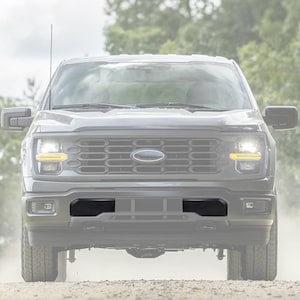 May include: Front view of a grey pickup truck with bright headlights and a black grill. The truck is driving on a dirt road with trees in the background. The Ford emblem is visible on the grill.