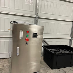 May include: A stainless steel industrial machine with a digital display, red and green buttons, and a flow meter. A black plastic container is next to the machine. The machine is on a concrete floor.