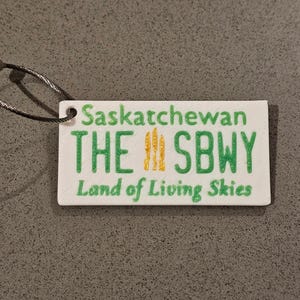 Może przedstawiać: Prostokątny biały brelok do kluczy z napisem "Saskatchewan THE SBWY Land of Living Skies" w kolorze zielonym i żółtym. Brelok ma metalową pętlę.