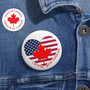 Peut inclure: Un bouton blanc avec une feuille d'érable rouge et le texte "I Stand With Canada" sur un fond en forme de cœur avec les drapeaux américain et canadien. Le bouton est sur une veste en jean bleue.