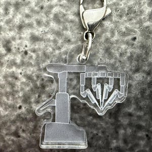May include: A clear acrylic charm with a silver-colored clasp. The charm features a stylized, geometric design in a light gray color. The design appears to be a logo or emblem, possibly for a brand or organization.