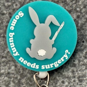 May include: A teal badge reel with a gray bunny holding a scalpel. The text "Some bunny needs surgery?" is in white around the edge of the badge. The badge reel has a white plastic clip and a metal retractable cord.
