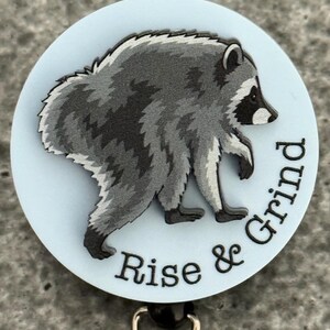 Peut inclure: Un enrouleur de badge circulaire avec un fond bleu clair et une illustration grise et noire d'un raton laveur en marche. Le texte "Rise & Grind" est imprim&eacute; en noir le long de la courbe inf&eacute;rieure, avec un clip m&eacute;tallique &agrave; la base.