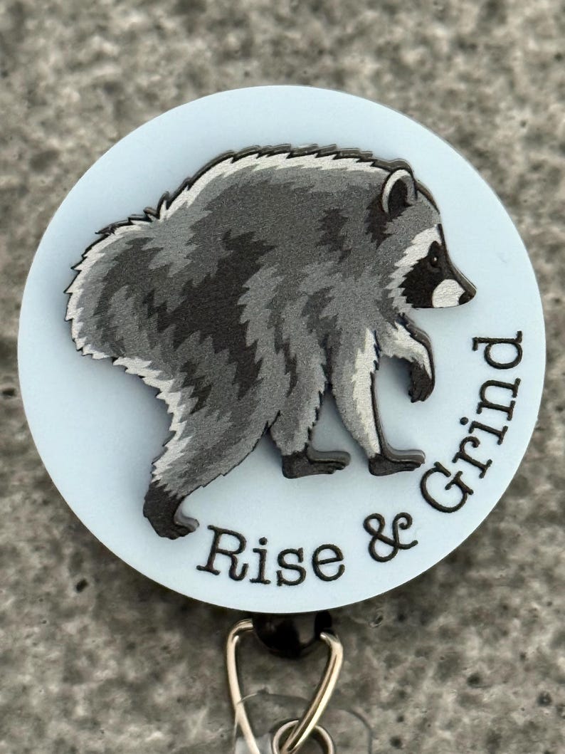 Peut inclure: Un enrouleur de badge circulaire avec un fond bleu clair et une illustration grise et noire d'un raton laveur en marche. Le texte "Rise & Grind" est imprim&eacute; en noir le long de la courbe inf&eacute;rieure, avec un clip m&eacute;tallique &agrave; la base.