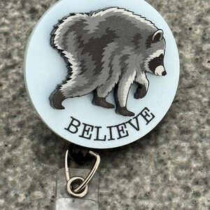 Peut inclure: Un enrouleur de badge circulaire avec un fond bleu clair et une illustration de raton laveur gris et noir. Le mot BELIEVE est imprim&eacute; en lettres capitales noires sous le raton laveur. Un clip en m&eacute;tal argent&eacute; et une sangle en plastique sont fix&eacute;s.