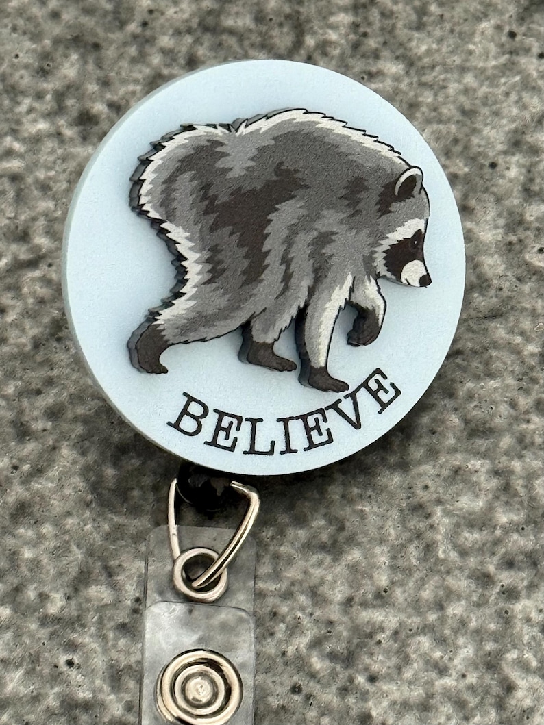 Peut inclure: Un enrouleur de badge circulaire avec un fond bleu clair et une illustration de raton laveur gris et noir. Le mot BELIEVE est imprim&eacute; en lettres capitales noires sous le raton laveur. Un clip en m&eacute;tal argent&eacute; et une sangle en plastique sont fix&eacute;s.