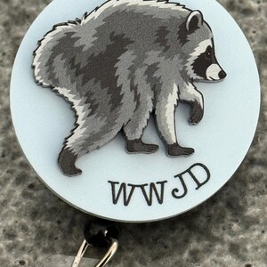 Peut inclure: Un enrouleur de badge circulaire avec un fond bleu clair et une illustration graphique d'un raton laveur gris et noir en marche. Sous le raton laveur, les lettres WWJD sont imprim&eacute;es dans une police noire simple. L'enrouleur a un clip en m&eacute;tal.