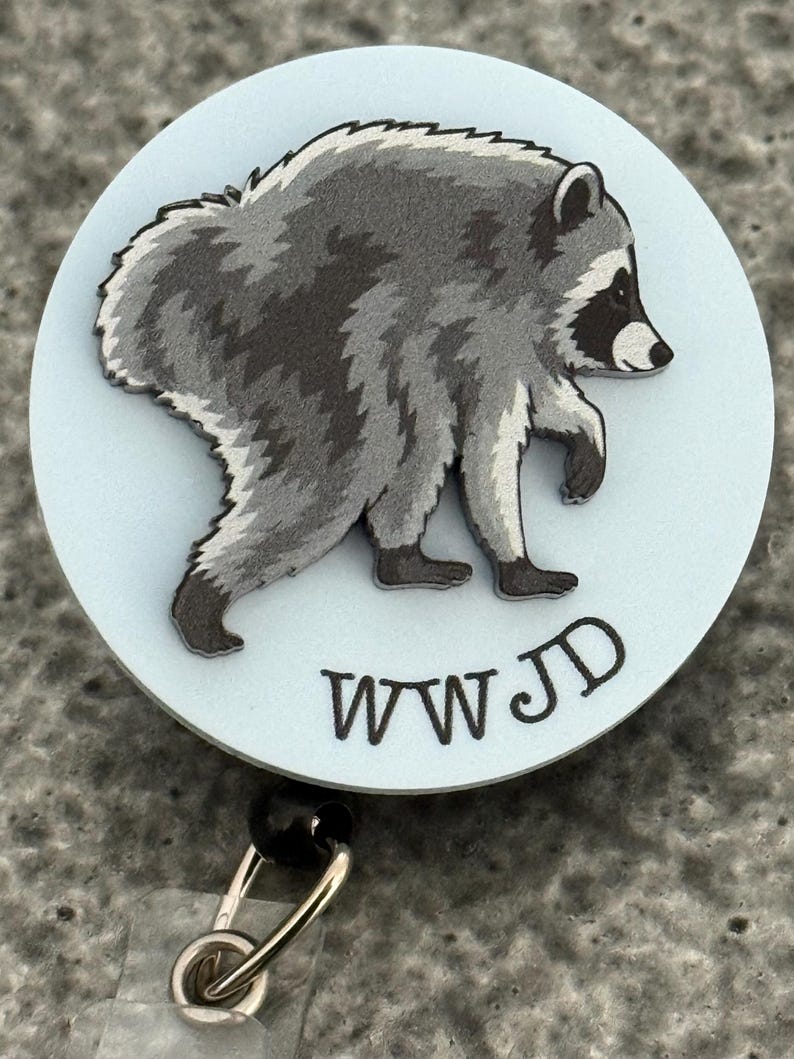 Peut inclure: Un enrouleur de badge circulaire avec un fond bleu clair et une illustration graphique d'un raton laveur gris et noir en marche. Sous le raton laveur, les lettres WWJD sont imprim&eacute;es dans une police noire simple. L'enrouleur a un clip en m&eacute;tal.