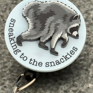 Peut inclure: Un enrouleur de badge rond avec un fond bleu clair et une illustration graphique d'un raton laveur gris et noir. Le texte "sneaking to the snackies" est imprim&eacute; en noir le long de la courbe inf&eacute;rieure de la face du badge rond.