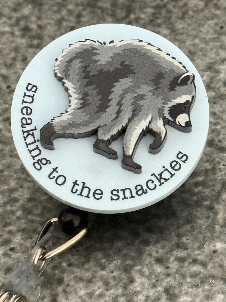 Peut inclure: Un enrouleur de badge rond avec un fond bleu clair et une illustration graphique d'un raton laveur gris et noir. Le texte "sneaking to the snackies" est imprim&eacute; en noir le long de la courbe inf&eacute;rieure de la face du badge rond.