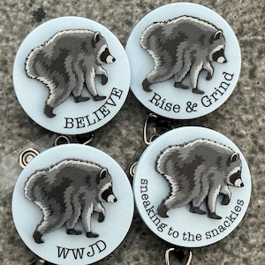 Peut inclure: Quatre enrouleurs de badge circulaires avec un fond bleu clair et une illustration de raton laveur gris. Chaque enrouleur affiche un texte noir unique : BELIEVE, Rise & Grind, WWJD et sneaking to the snackies, dispos&eacute; en bas ou sur le c&ocirc;t&eacute;.