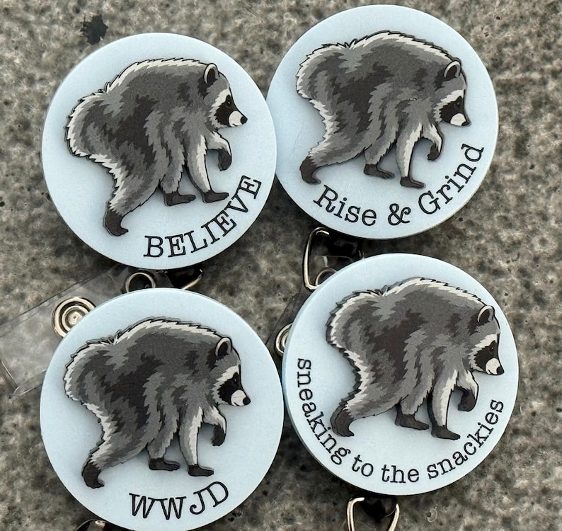 Peut inclure: Quatre enrouleurs de badge circulaires avec un fond bleu clair et une illustration de raton laveur gris. Chaque enrouleur affiche un texte noir unique : BELIEVE, Rise & Grind, WWJD et sneaking to the snackies, dispos&eacute; en bas ou sur le c&ocirc;t&eacute;.