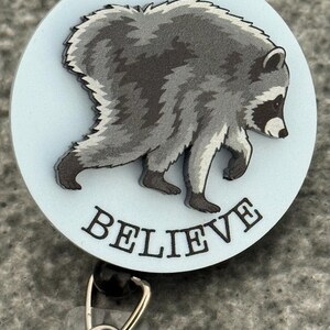 Peut inclure: Un enrouleur de badge circulaire avec un fond bleu clair et une illustration grise et noire d'un raton laveur en marche. Le mot BELIEVE est imprim&eacute; en lettres capitales noires sous le raton laveur. Un clip en m&eacute;tal argent&eacute; est fix&eacute; en bas.