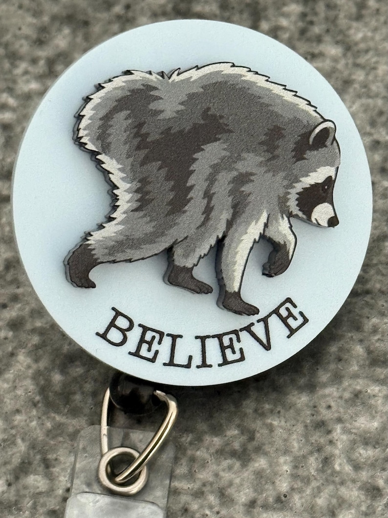 Peut inclure: Un enrouleur de badge circulaire avec un fond bleu clair et une illustration grise et noire d'un raton laveur en marche. Le mot BELIEVE est imprim&eacute; en lettres capitales noires sous le raton laveur. Un clip en m&eacute;tal argent&eacute; est fix&eacute; en bas.
