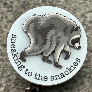 Peut inclure: Un enrouleur de badge circulaire avec un fond bleu clair et une illustration grise et noire d'un raton laveur en marche. Le texte "sneaking to the snackies" est imprim&eacute; en noir, en minuscules, avec une police sans empattement.