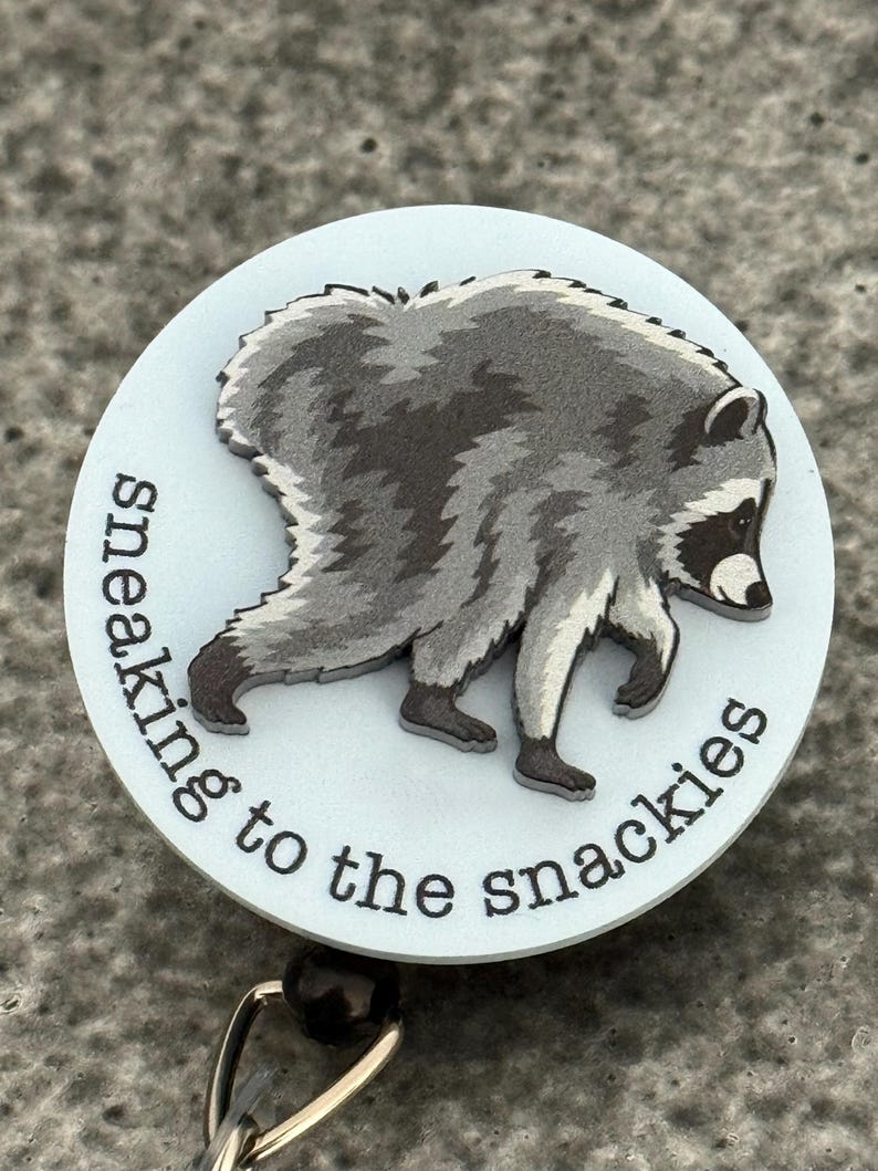 Peut inclure: Un enrouleur de badge circulaire avec un fond bleu clair et une illustration grise et noire d'un raton laveur en marche. Le texte "sneaking to the snackies" est imprim&eacute; en noir, en minuscules, avec une police sans empattement.
