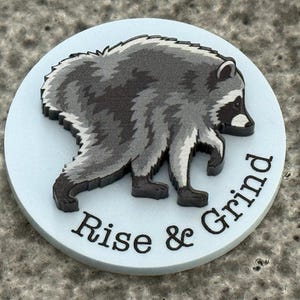 Peut inclure: Un disque circulaire bleu clair pr&eacute;sentant une illustration en relief d'un raton laveur gris fonc&eacute; et blanc en marche. Le texte "Rise & Grind" est imprim&eacute; en lettres capitales noires sans empattement le long de la courbe inf&eacute;rieure.