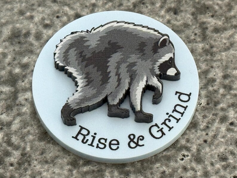 Peut inclure: Un disque circulaire bleu clair pr&eacute;sentant une illustration en relief d'un raton laveur gris fonc&eacute; et blanc en marche. Le texte "Rise & Grind" est imprim&eacute; en lettres capitales noires sans empattement le long de la courbe inf&eacute;rieure.