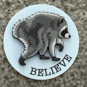 Peut inclure: Un badge circulaire bleu clair orn&eacute; d'une illustration d&eacute;taill&eacute;e d'un raton laveur gris et noir marchant vers la droite. Le mot BELIEVE est imprim&eacute; en lettres capitales noires et grasses le long de la courbe inf&eacute;rieure du badge circulaire.