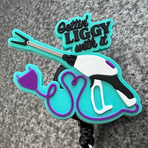 Puede incluir: Un portagafete retráctil azul turquesa con un cordón morado y un diseño blanco y negro de una pistola con el texto "Gettin' Liggy with it" en la parte superior.