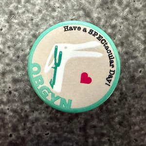 May include: A round button with a teal border and a beige center. The center features a white illustration of a medical tool, a red heart, and the text "OBGYN" and "Have a Spectacular Day!" in teal and black.