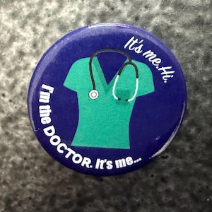 May include: A round, navy blue button with the text "I'm the DOCTOR. It's me..." in white. The button features a teal green medical scrub top and a white and black stethoscope graphic. The text "It's me. Hi." is also present.