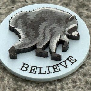 Peut inclure: Un jeton circulaire bleu clair pr&eacute;sentant une illustration gris fonc&eacute; et noire d'un raton laveur de profil. Le mot BELIEVE est imprim&eacute; en lettres capitales noires le long du bord inf&eacute;rieur du jeton, sous le graphique du raton laveur.