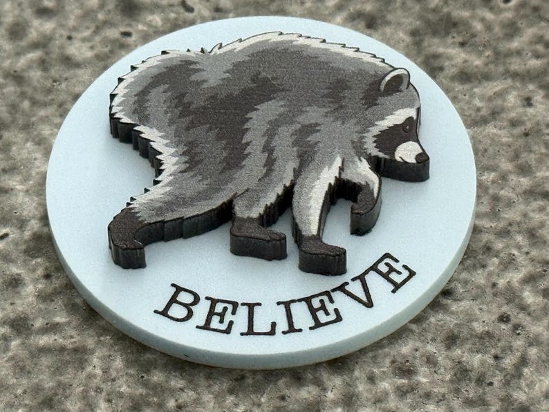Peut inclure: Un jeton circulaire bleu clair pr&eacute;sentant une illustration gris fonc&eacute; et noire d'un raton laveur de profil. Le mot BELIEVE est imprim&eacute; en lettres capitales noires le long du bord inf&eacute;rieur du jeton, sous le graphique du raton laveur.