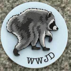 Peut inclure: Une plaque circulaire bleu clair pr&eacute;sentant une illustration en relief d'un raton laveur gris et noir marchant vers la droite. Les lettres WWJD sont imprim&eacute;es en noir au bas de la surface circulaire, positionn&eacute;es sous la figure du raton laveur.