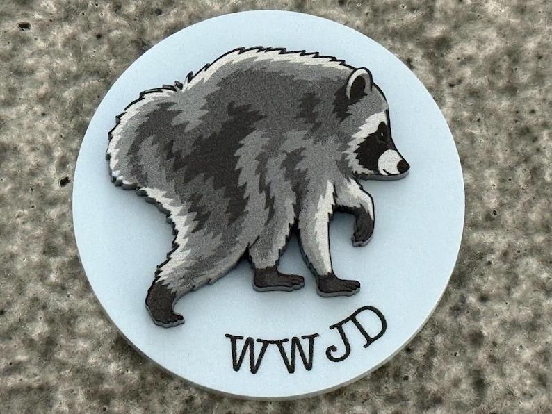 Peut inclure: Une plaque circulaire bleu clair pr&eacute;sentant une illustration en relief d'un raton laveur gris et noir marchant vers la droite. Les lettres WWJD sont imprim&eacute;es en noir au bas de la surface circulaire, positionn&eacute;es sous la figure du raton laveur.