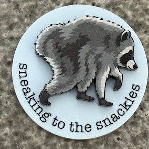 Peut inclure: Un autocollant circulaire blanc pr&eacute;sentant une illustration grise et noire d'un raton laveur qui marche. Le texte "sneaking to the snackies" est imprim&eacute; en noir, en minuscules, avec une police sans empattement le long du bord inf&eacute;rieur.