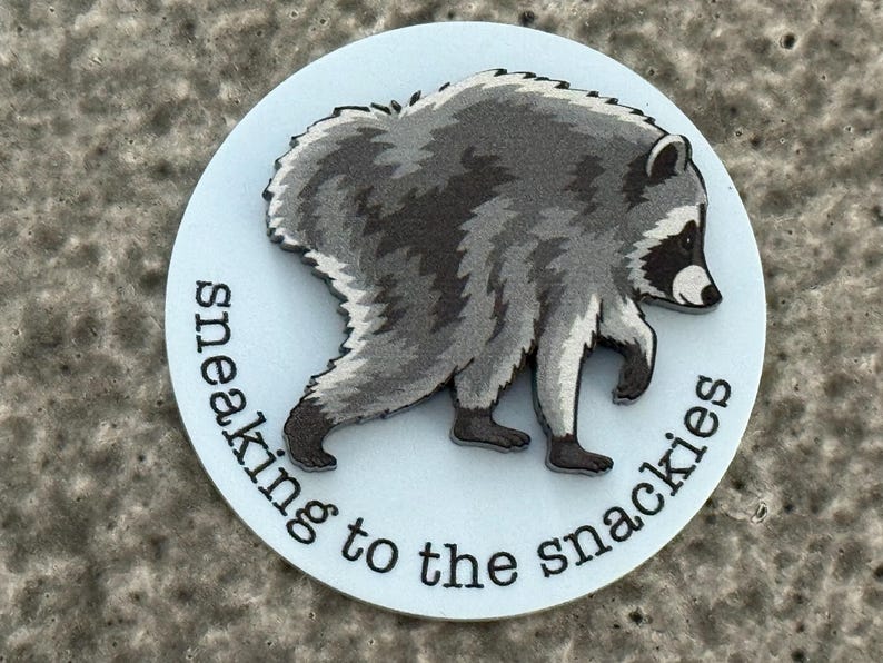 Peut inclure: Un autocollant circulaire blanc pr&eacute;sentant une illustration grise et noire d'un raton laveur qui marche. Le texte "sneaking to the snackies" est imprim&eacute; en noir, en minuscules, avec une police sans empattement le long du bord inf&eacute;rieur.