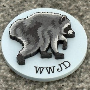 Peut inclure: Un disque circulaire bleu clair pr&eacute;sentant une illustration en relief d'un raton laveur dans des tons de gris, noir et blanc. Le raton laveur est repr&eacute;sent&eacute; de profil, marchant vers la droite. Le texte noir WWJD est imprim&eacute; sous le raton laveur.