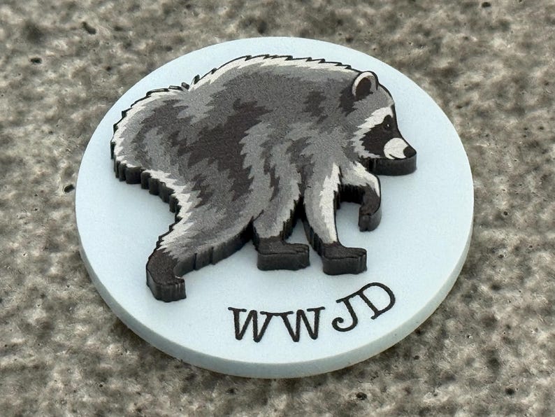 Peut inclure: Un disque circulaire bleu clair pr&eacute;sentant une illustration en relief d'un raton laveur dans des tons de gris, noir et blanc. Le raton laveur est repr&eacute;sent&eacute; de profil, marchant vers la droite. Le texte noir WWJD est imprim&eacute; sous le raton laveur.