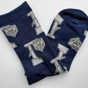 Peut inclure: Une paire de chaussettes bleu marine avec un motif répété de derricks pétroliers gris pixélisés et d'une chaîne de montagnes grises pixélisées. Les chaussettes sont faites d'une matière douce et confortable.